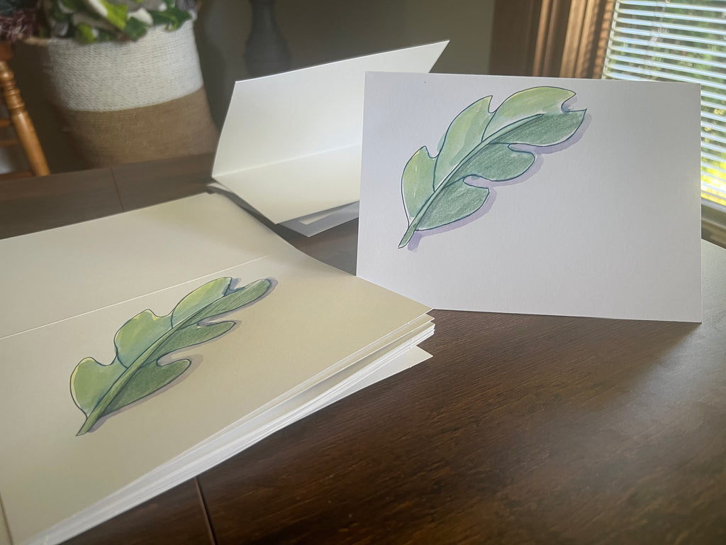 A stack of white greeting cards with hand-drawn green leaf illustrations, with one card standing upright on a wooden table in a home setting.