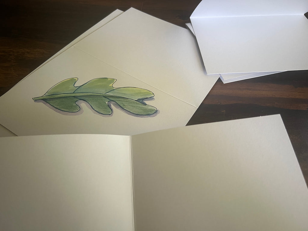 An open cream-colored notebook or card album on a dark wooden table, featuring a hand-drawn green leaf illustration on one of the blank pages.