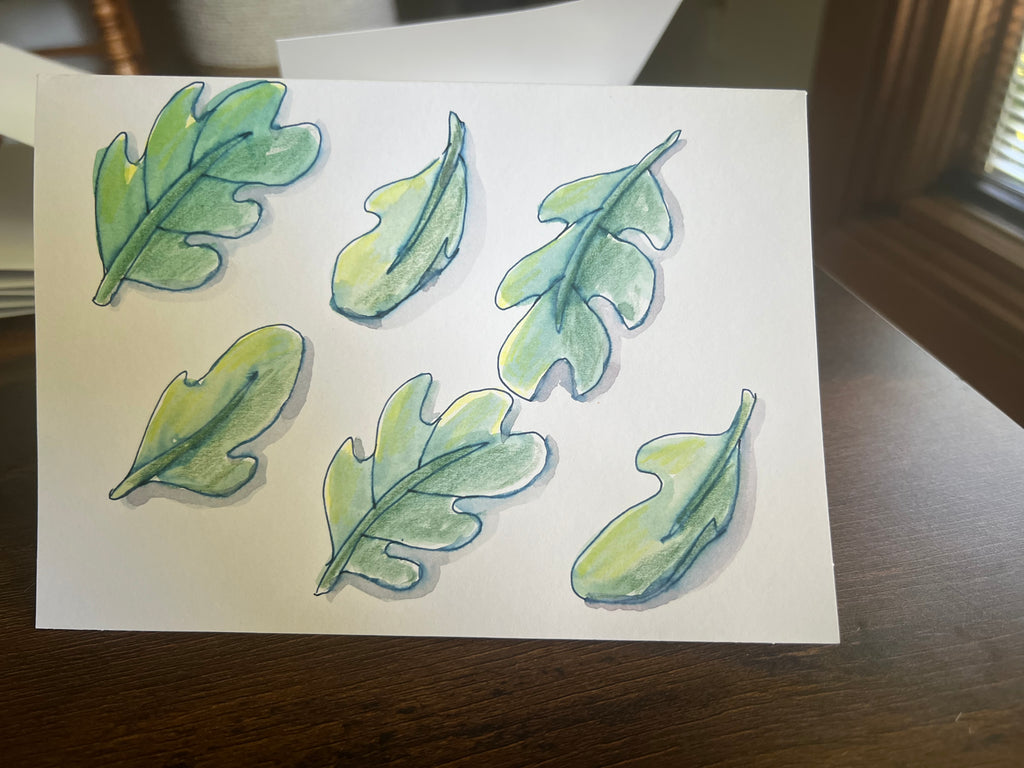 A watercolor painting of six green arugula leaves on a white sheet of paper, resting on a dark wooden table.