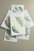 A stack of white art cards featuring watercolor illustrations of green leaves, arranged on a desk.